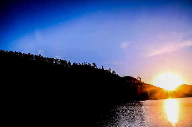 A beautiful sunset over a lake with a mountain in the background. The sky is a deep blue color with a few clouds. The sun is setting and its reflection can be seen on the water