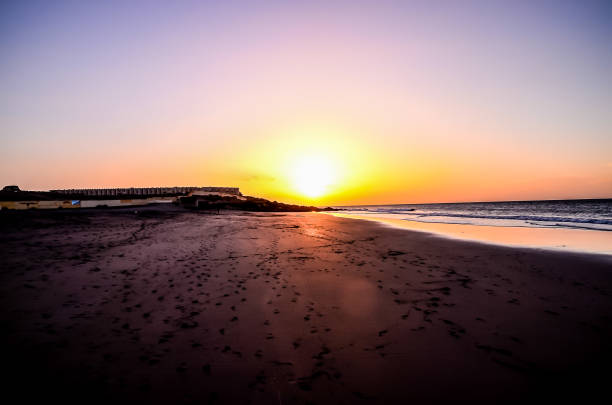 A beach with a sunset in the background. The sky is a mix of purple and orange. The beach is empty, with no people visible
