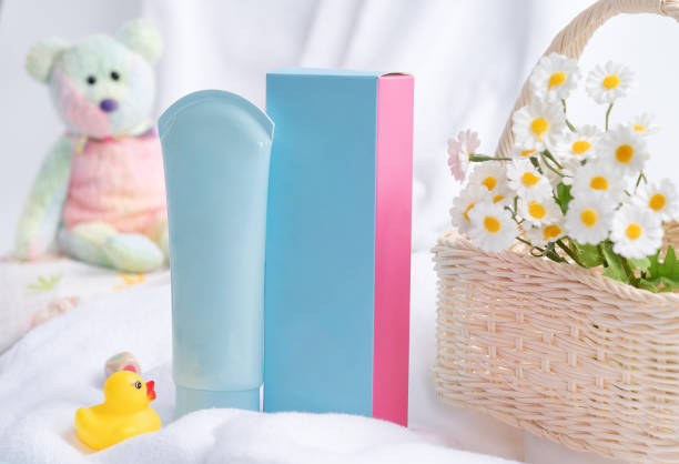 The image features a soft, light-colored setting with various child-related items. There is a plush teddy bear in pastel colors, a blue container, a pink block, and a yellow rubber duck. Additionally, a wicker basket is present, filled with small daisies, alongside a white towel and some colorful block pieces.