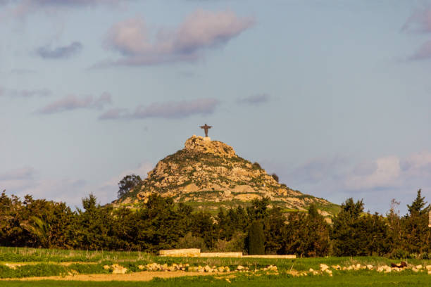 Large metal cross standing proudly on a hill, surrounded by lush vegetation, under a bright blue sky dotted with fluffy clouds, creating a serene and picturesque landscape