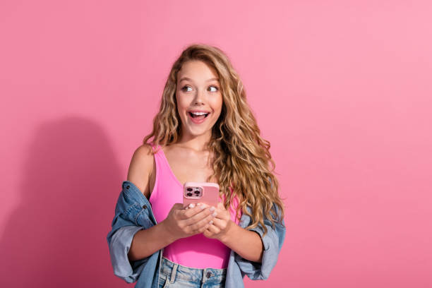 istock Young woman expressing joy while using smartphone against a pink background in a stylish casual outfit 2192776608