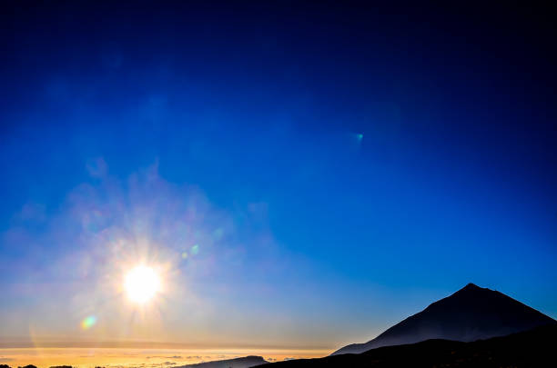 A mountain is silhouetted against a blue sky. The sun is shining brightly, casting a warm glow over the scene. The mountain is covered in snow, and the sky is clear and cloudless