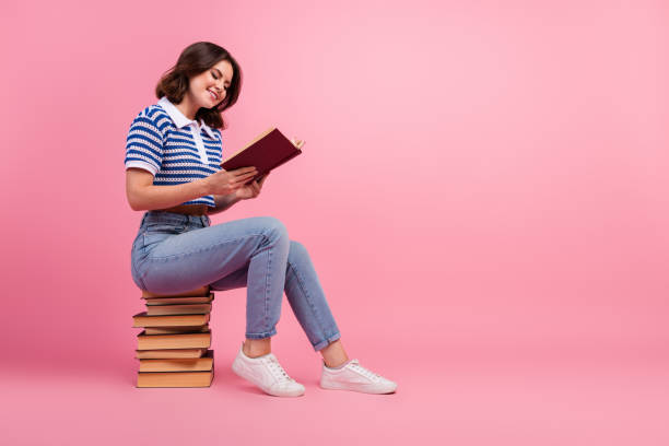 istock Young brunette woman reading a book while sitting on a stack of books against a pink background, wearing a striped knitwear polo 2192781051