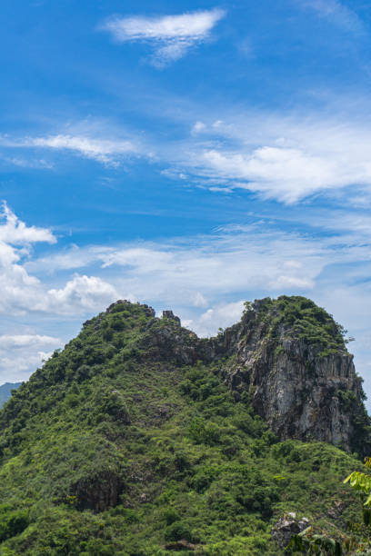 A lush, green mountain peak stands prominently against a bright blue sky dotted with white clouds.