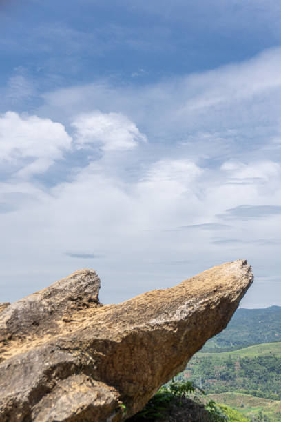 A rocky outcrop extends outward against a blue sky with white clouds, overlooking a green, hilly landscape.