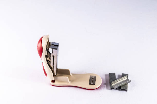A red and white stapler with a silver clip is sitting on a white surface. The stapler is open and the clip is missing. There are several small metal staples scattered around the stapler