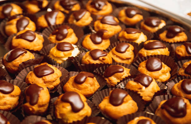 A tempting assortment of golden cream puffs topped with rich, glossy chocolate glaze, beautifully arranged in individual wrappers. These decadent pastries are perfect for dessert tables, celebrations, or as a delightful treat for chocolate lovers. The close-up view showcases their fluffy texture and indulgent toppings, creating a feast for the eyes and the palate