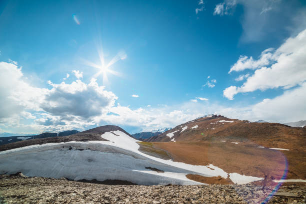 Scenic view to sunlit high alpine valley between glacier and sharp spiky rocks on hill against large snowy peak far away under bright sun among clouds in blue sky. Big snow-capped thorny mountain top.