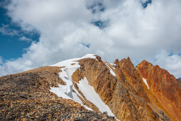 istock Snow-capped mountain ridge with cornice and red sharp rocks on top in sunlight. Snow cap and shiny pointy peak of gold color. Snow dome and colorful rocky peaked top. Sunny cloudy high mountains view. 2192806838