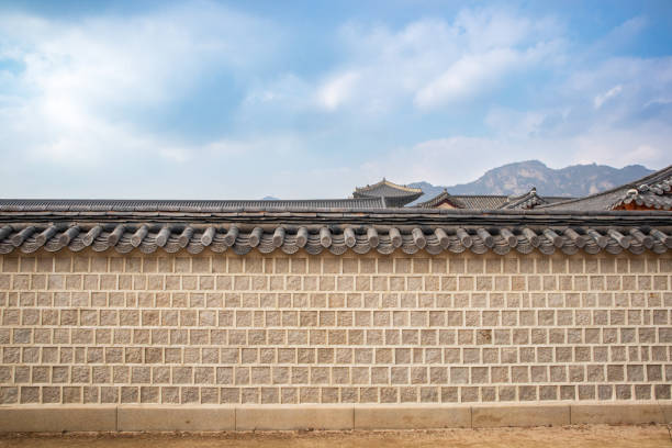 A traditional Korean wall features stone and wooden elements, reflecting the heritage of the historical district. The backdrop includes intricate roof designs under a clear sky.