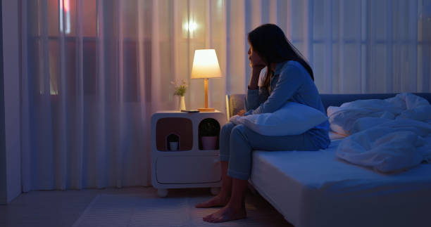 side view asian woman sits alone by the bedside in a dark room feeling troubled and unable to sleep
