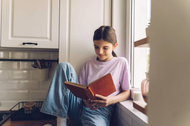 istock Young girl reading book by window in cozy kitchen 2192819757
