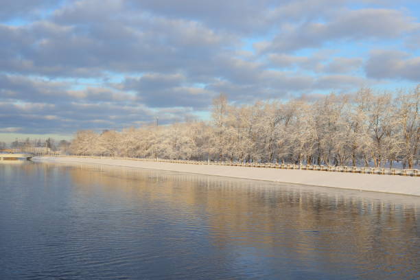 A beautifully picturesque winter scene showcases frosted trees lining a tranquil river beneath a bright, clear sky