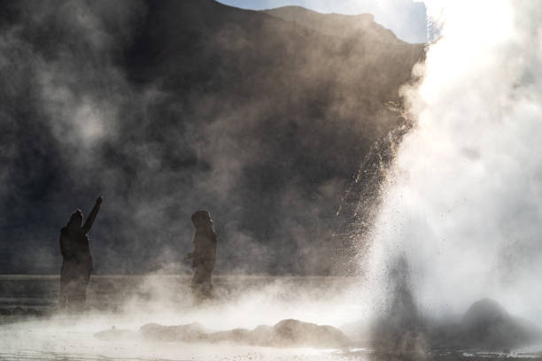 Atmospheric scenery featuring silhouetted figures among rising steam clouds from geysers in the Atacama Desert during early morning light.