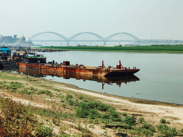 Tranquil riverside scene showcasing moored vessels with a scenic arched bridge in Myanmar.