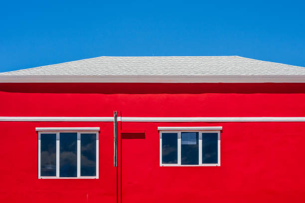 A striking image of a vibrant red building with white-framed windows and a clean white roof. The bold red contrasts beautifully with the cloudless bright blue sky, creating a minimalist yet visually captivating composition. This architectural detail emphasizes symmetry and bold color contrasts, making it a perfect representation of urban simplicity.