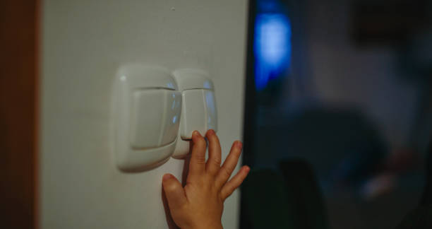 A Caucasian boy extends his hand to a light switch in a warmly lit home environment. The scene highlights the child's curiosity and the cozy interior setting.