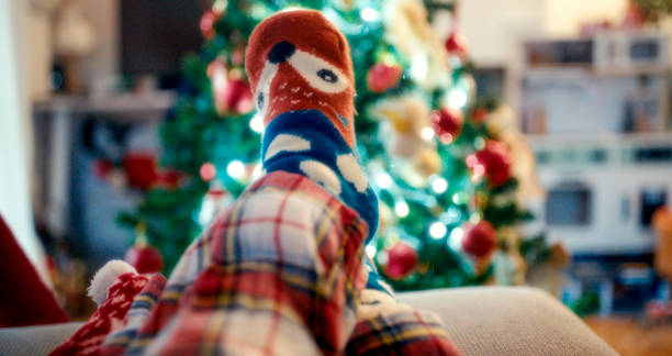 A person wearing festive Christmas socks, relaxing on a sofa. The background features a warmly lit Christmas tree adorned with colorful ornaments, creating a cozy holiday atmosphere at home.