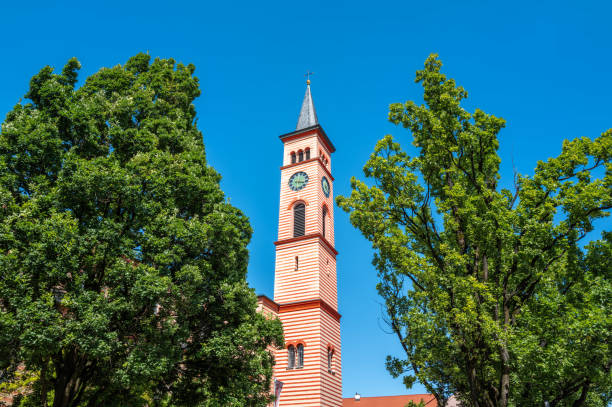 A tall red and white striped church tower with a clock in Friedberg, surrounded by green trees against a clear blue sky.