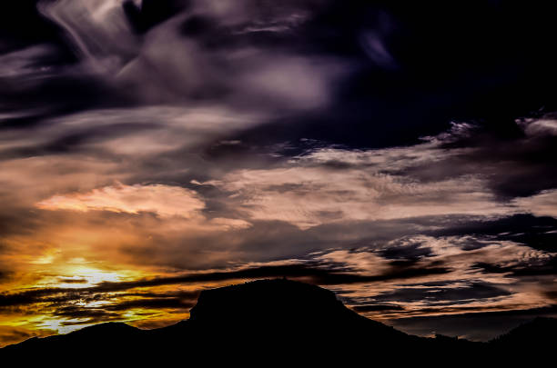A mountain range is silhouetted against a dark sky with a bright orange sun. The sky is filled with clouds, giving the scene a moody and dramatic feel