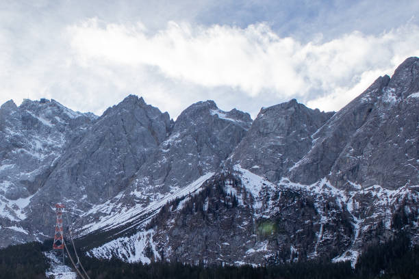 Rocky mountain peaks tower majestically, covered in snow and framed by clouds, while a cable car ascends along the slope, offering breathtaking views of the terrain.