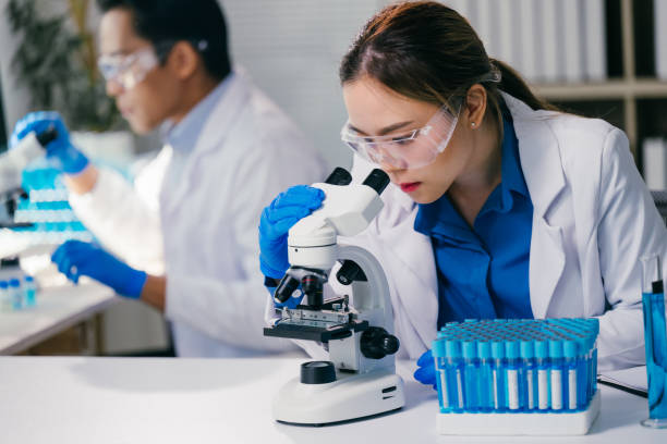 istock Young female scientist wearing gloves and safety glasses looking into a microscope, analyzing samples in test tubes, with a male scientist working in the background 2192902500