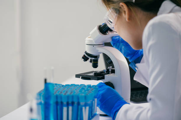 istock Scientist wearing a lab coat and gloves is carefully analyzing a sample under a microscope, surrounded by test tubes containing blue liquid, in a brightly lit laboratory setting 2192902535