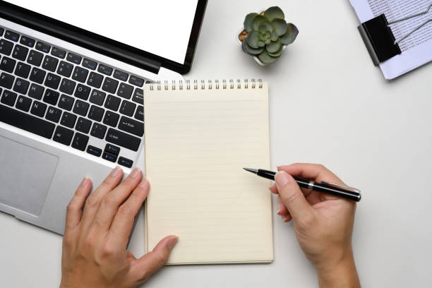 istock Above view of man writing in a spiral notebook while working on a laptop at white desk 2192911019