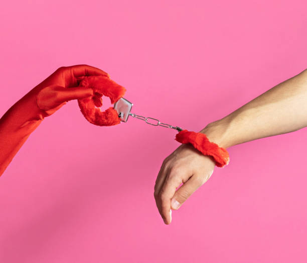 Woman's and a man's hand clasped by red fluffy handcuffs on a pink background stock photo