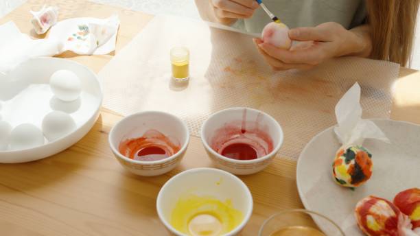 istock A Woman Paints Easter Eggs at a Sunlit Table, Surrounded by Bowls of Dye and White Eggs. 2192919715