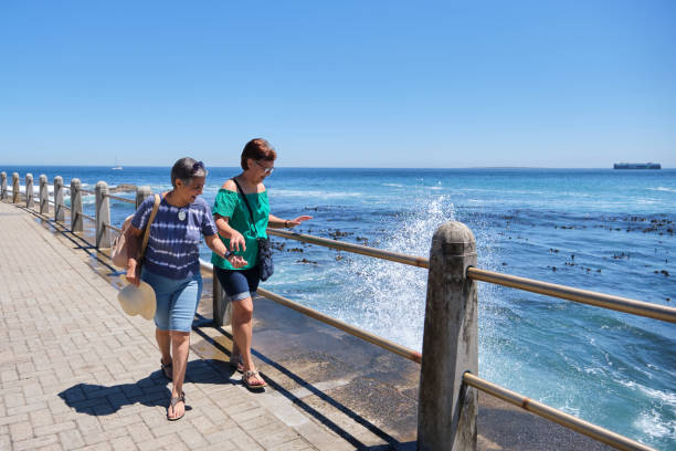 Two senior mixed race friends explore their home city while on vacation. They are walking along the Seapoint Promenade enjoying the views over the sea as the waves splash