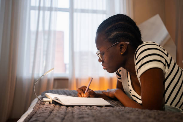 istock Young black woman lying on bed writing in notebook by lamp light 2192929596