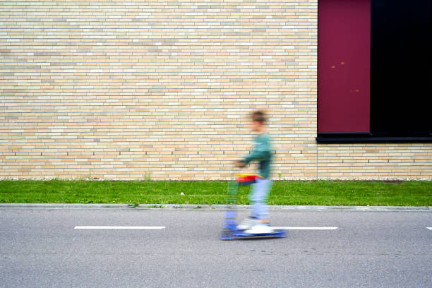 A child on a scooter speeds past a brick wall, blurring motion against static urban backdrop.