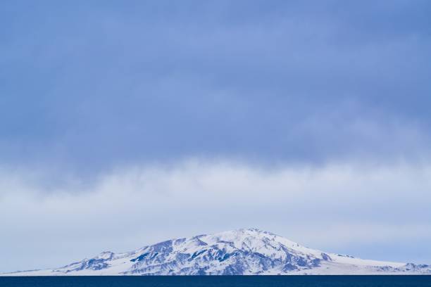 A serene snow-capped mountain under a vast blue sky with a tranquil sea in the foreground.