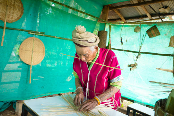 Concentrated mature thai man is doing traditional bamboo craft under sun protection in rural village working outdoors in a small village in remote location in Chiang Mai province. Man with wrinkled face  is making bamboo baskets . He  is wearing traditional northern local thai clothes and is working concentrated.   His workplace with table is under a wooden framework with wooden ceiling made from small trees.  Two side are covered with a green canvas as sun protection which is coming backside of the man into the village and workplace in mountain area. Workplace with table is in open area  of village, no indoors space, 100% outdoors space under wooden framework. People of village are also making traditional thai clothes and textiles with loom outside below the stilt houses in front of several houses. Scene is in area of mountains of ban Pong