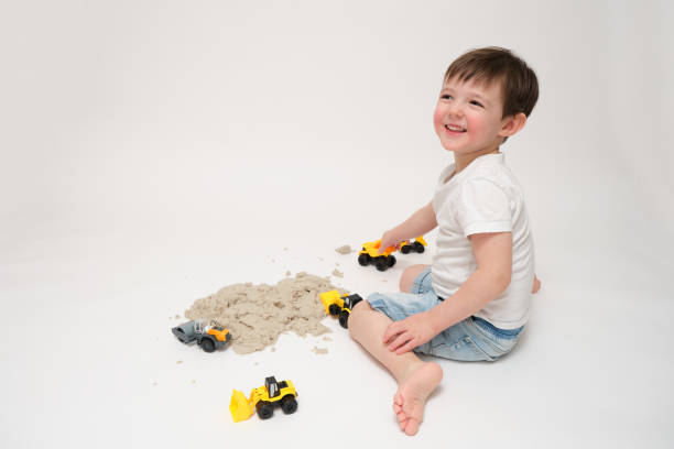 A toddler baby is happily playing with kinetic sand on a white studio background. The smiling boy uses his hands to shape toys out of kinetic sand indoors. Kid is three years old