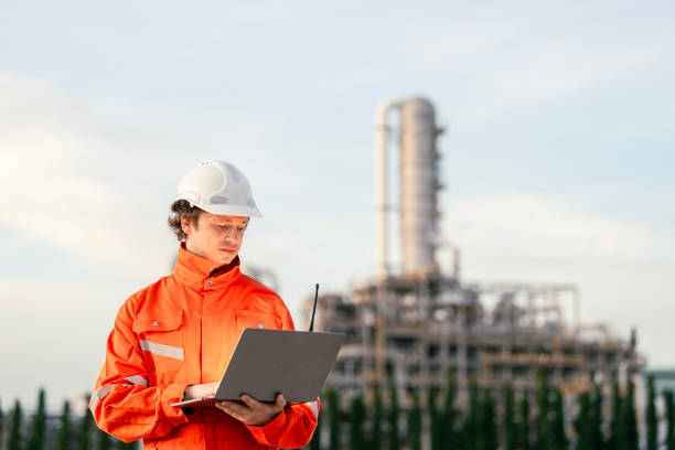 istock Engineer in bright orange reflective suit with safety helmet and laptop, standing in front of an industrial refinery, symbolizing leadership in technology, infrastructure, and safety management. 2192986251