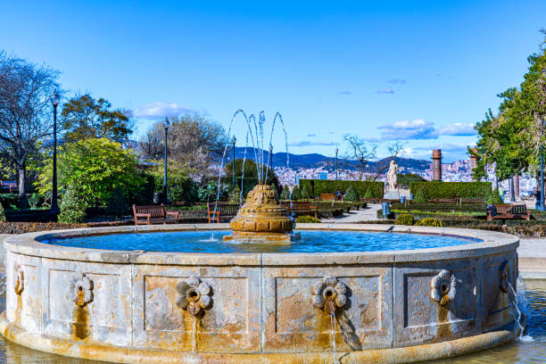 Stone fountain decorated with carved flowers, water jets coming out from central pipes, Joan Brossa Gardens on Montjuic, trees and cityscape in background, sunny day in Barcelona, Catalonia, Spain