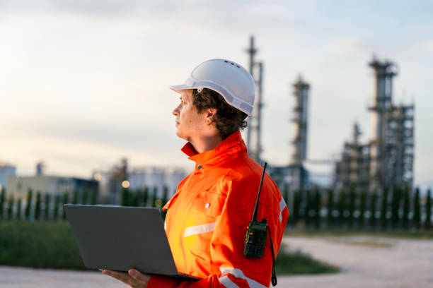 istock Engineer in bright orange reflective suit with safety helmet and laptop, standing in front of an industrial refinery, symbolizing leadership in technology, infrastructure, and safety management. 2192989159