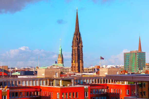 View of Hamburg's skyline featuring the spire of St. Nikolai Memorial and the green dome of Hamburg City Hall, surrounded by modern and traditional architecture under a bright blue sky