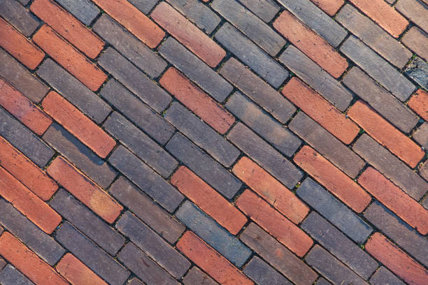 A close-up view of a herringbone patterned street surface in a Dutch village, made of red bricks. Weeds sprout between the bricks, adding a touch of nature to the structured paving. This intricate brickwork design showcases the traditional aesthetic and durability of Dutch village roads.