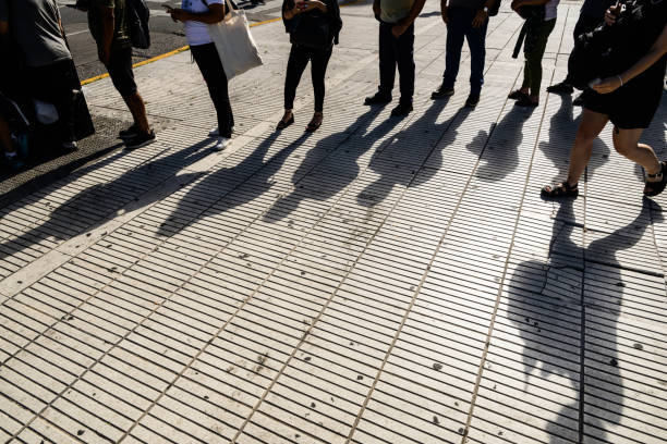 A diverse group of individuals is gathered on a bustling sidewalk, casting their long shadows onto the sunlit ground beneath them