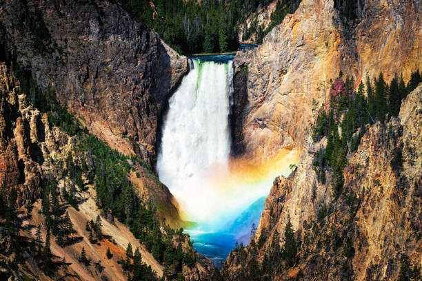 The image showcases a dramatic waterfall cascading over rocky cliffs, surrounded by dense greenery and an arched rainbow. The photograph highlights the natural beauty and dynamic landscapes found in scenic parks.