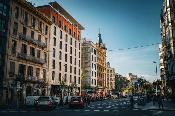 Barcelona, Spain - Nov 14 2024: A view of a street during the sunset. Some cars are stopped at the gtraffic light and some pedestrians are at the crosswalk. Buildings and trees are visible.