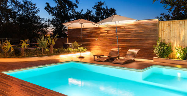 istock Modern backyard with illuminated swimming pool and lounge chairs under umbrellas. Luxury outdoor space surrounded by greenery and warm lighting 2193033588
