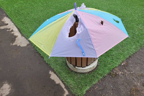 Torn multicolored umbrella covering a public trash bin on a grass and pavement background. Outdoor urban scene.