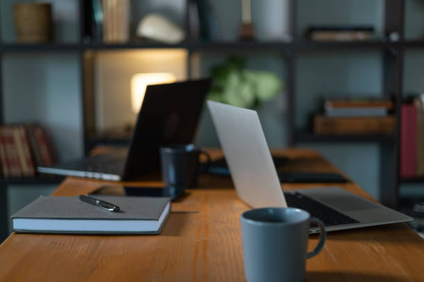 Cozy home office setup featuring open laptops, a notebook, pens, and coffee mugs on a wooden desk, with a blurred bookshelf in the background, creating a warm and inviting workspace atmosphere