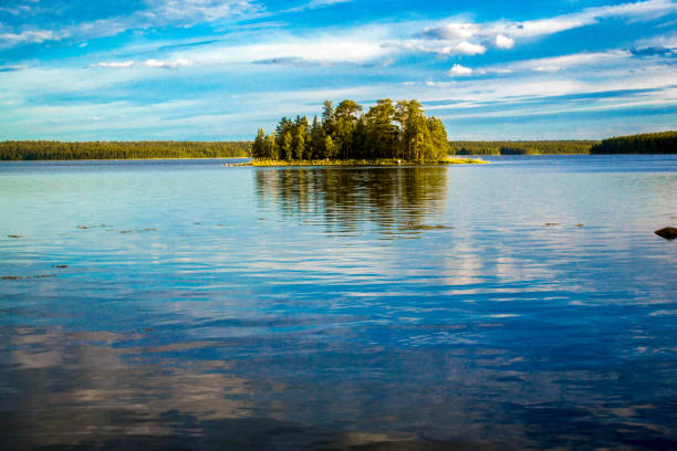 Lonely island covered with trees against a background of blue sky with clouds. The blue sky and clouds are reflected in the water. Small waves create soft reflections