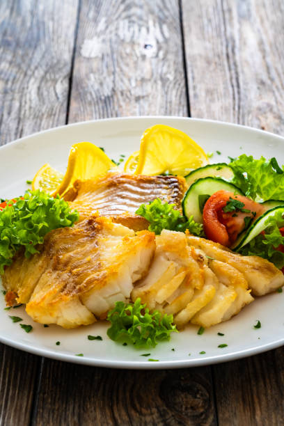 istock Fried codfish loin and fresh vegetables served on white plate on wooden table. 2193057081