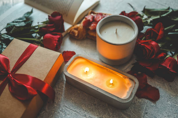 istock Concrete and ceramic candles, one glowing, create a cozy atmosphere with a kraft gift box tied with a red ribbon, red roses, petals, and a pastry. A romantic setting with a book in the background 2193064625